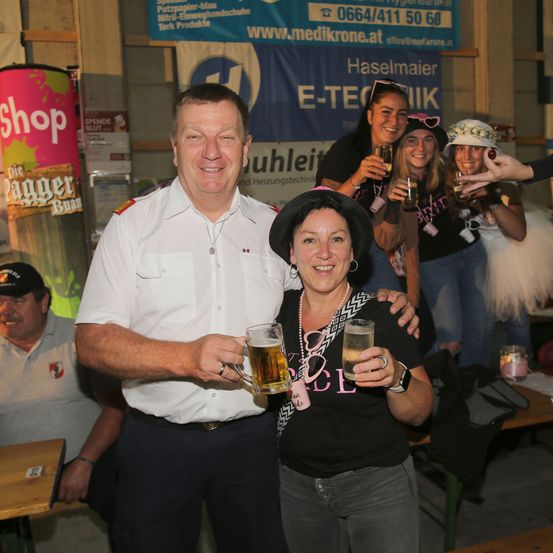 Ein Mann und eine Frau halten Biergläser und lächeln. Sie stehen nebeneinander. Hinter ihnen stehen drei Frauen mit Biergläsern. Sie sind wahrscheinlich auf einer Party.