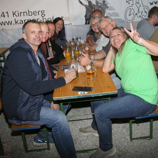 Eine Gruppe von Menschen sitzt an einem Tisch und hält Biergläser in den Händen. Eine Frau lächelt und macht ein Friedenszeichen. Hinter ihnen ist eine Wand mit einem Banner, auf dem '41 Kirnberg' steht.