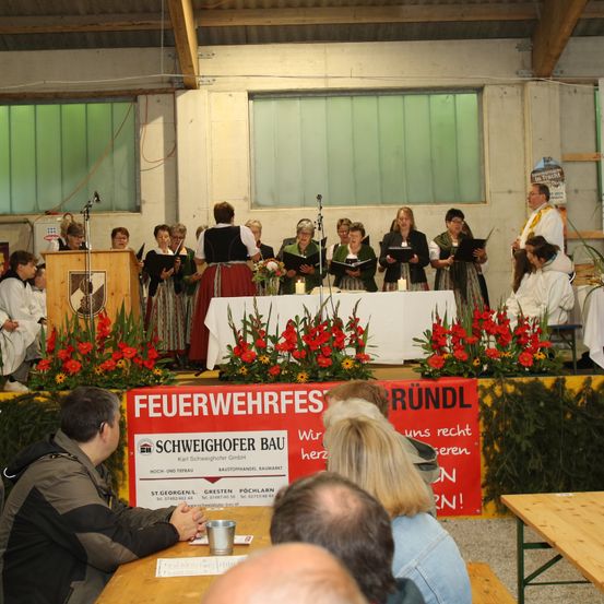 Ein Chor von Menschen in traditioneller Kleidung tritt vor einer Bühne mit roten Blumen auf. Ein Banner auf der Bühne heißt 'Feuerwehrfest Schweighofer Bau'. Ein Publikum sitzt vor der Bühne.