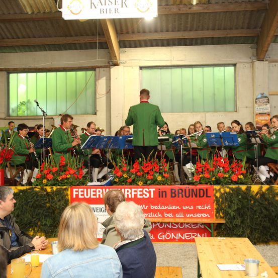 Ein Blasorchester spielt auf der Bühne vor roten Blumen. Ein Publikum sitzt davor. Ein Banner mit der Aufschrift 'Feuerwerkfest in Brundl'.