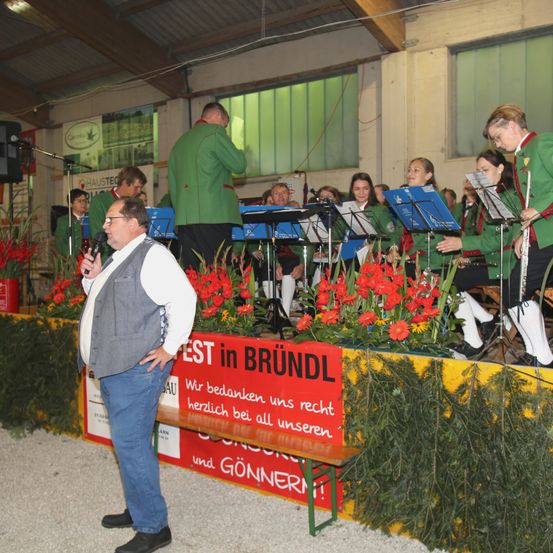 Ein Mann steht mit einem Mikrofon vor einer Gruppe von Musikern auf der Bühne. Sie sind in grünen Uniformen gekleidet. Hinter ihnen steht ein Schild mit der Aufschrift 'Fest in Brundl'. Auf der Bühne befinden sich Blumen und Grünpflanzen.