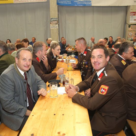 Eine Gruppe uniformierter Personen sitzt an einem langen Tisch in einem Saal, lächelt und interagiert miteinander. Sie halten Gläser mit Getränken und einige tragen Krawatten.