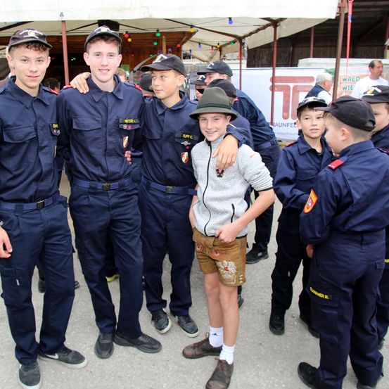 Mehrere junge Männer in Uniformen stehen in einer Reihe unter einem Zelt. Einer trägt einen Hut und ein graues Shirt, lächelt. Andere tragen Hüte und Uniformen mit Abzeichen. Ein verschwommener Hintergrund zeigt weitere Menschen und ein Banner.
