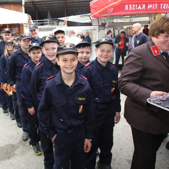 Eine Gruppe von Kindern in Uniformen steht in einer Reihe mit einem lächelnden Erwachsenen an der Seite. Sie nehmen möglicherweise an einer Parade oder einem Event teil. Dahinter befindet sich ein Stand mit einem roten Vordach.