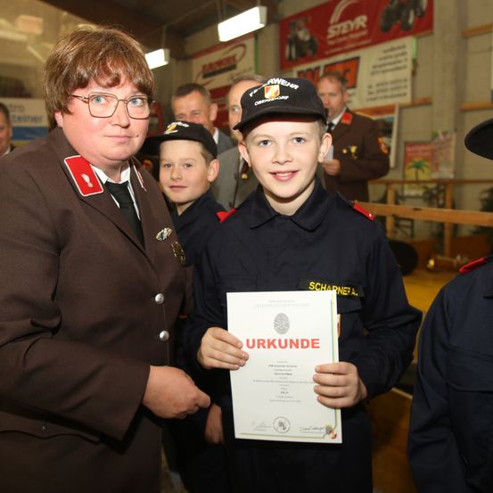 Eine Frau in Uniform steht mit einem lächelnden Jungen, der ein Zertifikat hält, umgeben von anderen uniformierten Kindern und Erwachsenen in einem Raum.