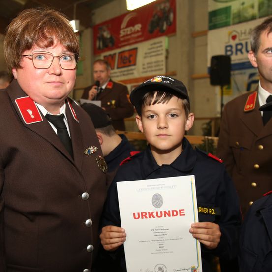 Ein junger Junge in Uniform hält ein Zertifikat, neben ihm steht eine Frau in Uniform. Ein Mann hält ein Mikrofon hinter ihnen, und andere in Uniform umgeben sie.