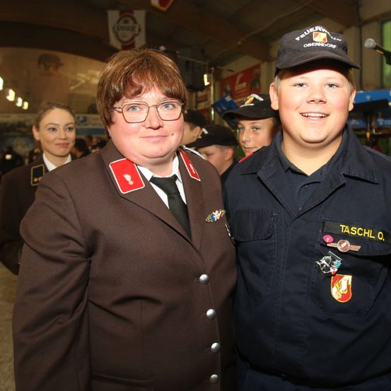 Zwei Menschen in Uniformen lächeln in die Kamera. Eine Frau mit Brille und ein Mann mit Hut. Sie sind wahrscheinlich bei einer Veranstaltung. Dahinter stehen weitere Menschen in Uniformen.