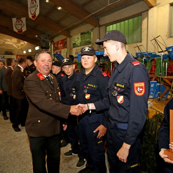 Ein Militäroffizier in Uniform gibt einem Soldaten in schwarzer Uniform die Hand. Dahinter stehen mehrere Soldaten in einer Reihe und tragen Mützen. Im Hintergrund befinden sich Banner und Musikinstrumente.
