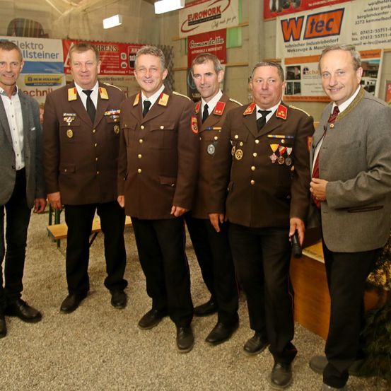 Fünf Männer in Uniformen, möglicherweise Militär, stehen in einer Reihe mit einem lächelnden Gesichtsausdruck. Dahinter befinden sich Banner mit Text und Logos. Der Boden ist mit Teppich bedeckt.