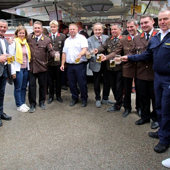 Eine Gruppe von Menschen, darunter Männer in Militäruniformen und eine Frau mit einem gelben Schal, stehen zusammen und halten Biergläser. Sie lächeln und posieren für ein Foto.