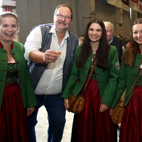 Ein Mann und drei Frauen stehen zusammen und lächeln. Der Mann hält ein Getränk in der Hand. Die Frauen tragen grüne Jacken und rote Röcke.