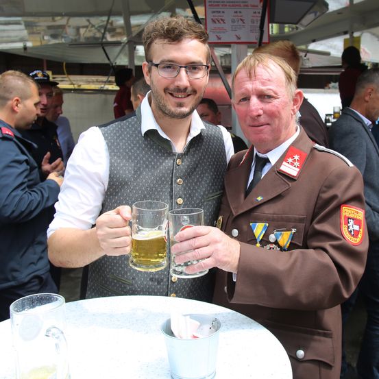 Zwei Männer, einer in Uniform und einer in einem Weste, stehen bei einem Tisch mit Bierkrügen. Dahinter stehen und sprechen andere Leute.