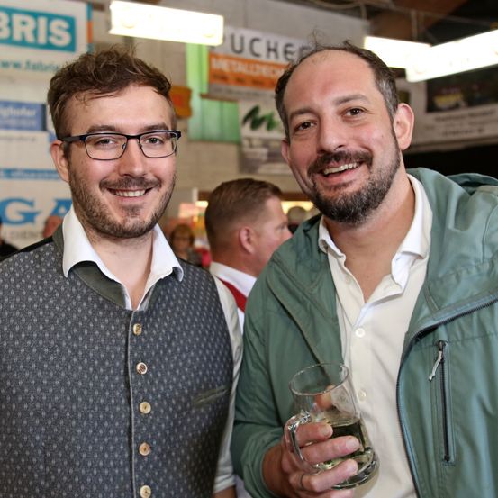 Zwei Männer lächeln und posieren für ein Foto. Der Mann auf der linken Seite trägt eine Brille und eine Weste, während der Mann auf der rechten Seite ein Glas mit einem Getränk in der Hand hält. Hinter ihnen ist ein weiterer Mann teilweise zu sehen.