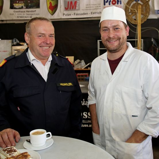 Zwei Männer stehen nebeneinander und lächeln, einer in einer Feuerwehruniform und der andere in einer weißen Kochjacke. Sie stehen vor einem Tisch mit Essen und einer Tasse Kaffee. Hinter ihnen ist ein Banner mit Logos und Text.