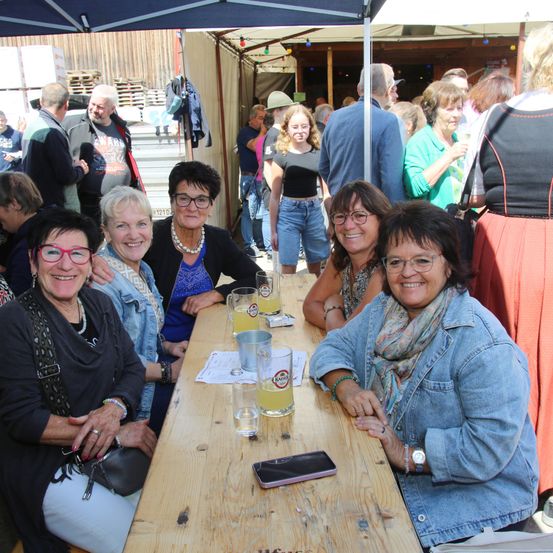 Eine Gruppe lächelnder Frauen mit Brillen sitzt um einen Tisch mit Getränken bei einer Veranstaltung im Freien, mit mehreren Personen und Ständen im Hintergrund.