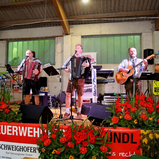 Drei Musiker treten auf einer Bühne mit Blumendekoration auf. Zwei spielen Akkordeon und einer Gitarre. Die Bühne ist mit Lautsprechern und einem Banner mit der Aufschrift 'Schweighofer' ausgestattet.