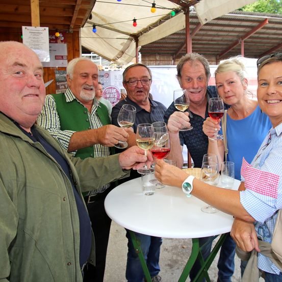 Fünf Personen versammeln sich um einen Tisch, lächeln und halten Weingläser in der Hand. Sie scheinen sich auf einem Weinverkostungsevent zu befinden, mit einem weißen Tisch und einem Schirm im Hintergrund.
