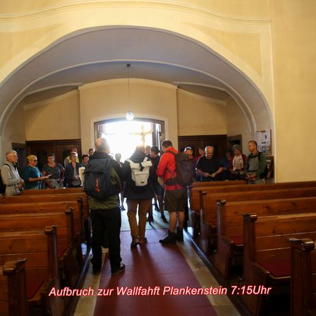 Eine Gruppe von Menschen mit Rucksäcken steht in einer Kirche. Die Kirche hat Holzbänke, einen Bogen und ein Fenster mit einem Kreuz. Der Text auf dem Bild lautet: 'Aufbruch zur Wallfahrt Plankenstein 7:15 Uhr.'