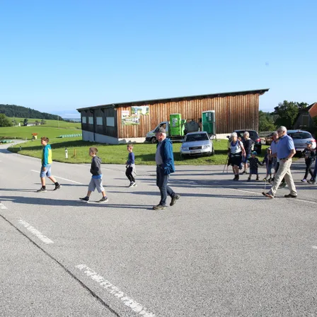 Eine Gruppe von Menschen geht auf einer Straße, an der Autos geparkt sind. Einige tragen Turnschuhe, während andere Gehstöcke haben. Ein Gebäude befindet sich auf der linken Seite und ein Rasenbereich auf der rechten Seite.