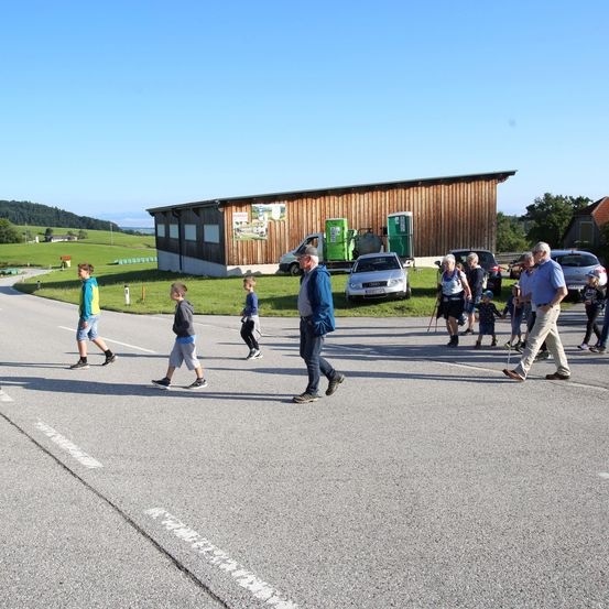 Eine Gruppe von Menschen geht auf einer Straße, an der Autos geparkt sind. Einige tragen Turnschuhe, während andere Gehstöcke haben. Ein Gebäude befindet sich auf der linken Seite und ein Rasenbereich auf der rechten Seite.