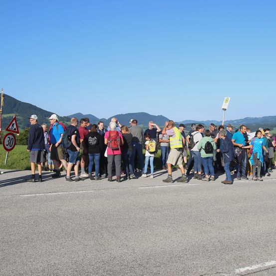 Eine Gruppe von Menschen steht am Straßenrand, wahrscheinlich auf etwas wartend. Sie sind alle für Outdoor-Aktivitäten gekleidet. Hinter ihnen befinden sich Berge und einige Schilder.