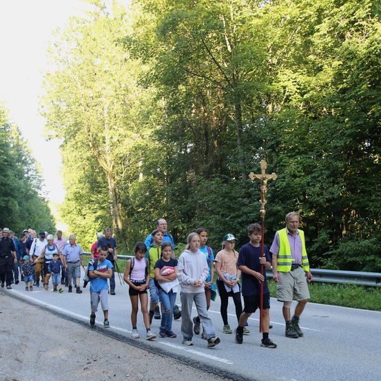 Eine Gruppe von Menschen geht auf einer Straße mit Bäumen auf beiden Seiten. Ein Mann in einer gelben Weste hält einen Stab mit einem Kreuz.