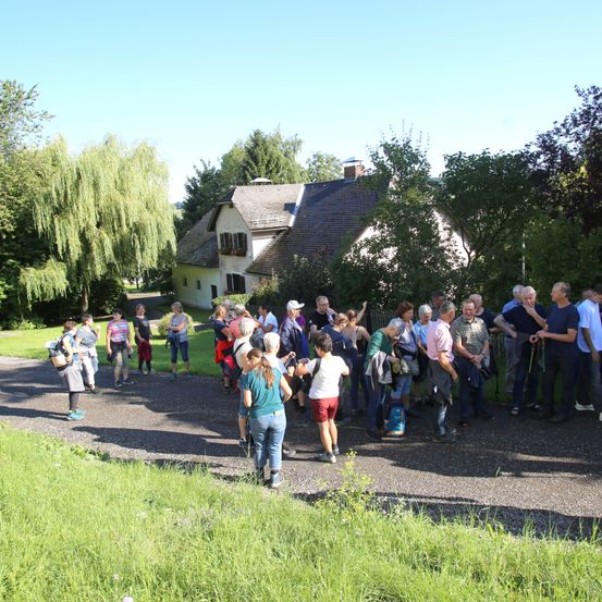 Eine Gruppe von Menschen steht auf einem Pfad vor einem Haus, wahrscheinlich auf einer Wanderung. Einige tragen Hüte und Turnschuhe, während andere Rucksäcke tragen. Das Haus hat ein graues Dach, und das Gebiet ist von Bäumen und Gras umgeben.