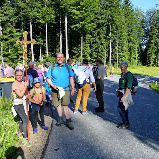 Eine Gruppe von Menschen steht auf einer Straße, ein Mann vorne mit einem Kreuz. Ein Mädchen hält ein Kreuz, während andere Rucksäcke tragen. Bäume umgeben sie.