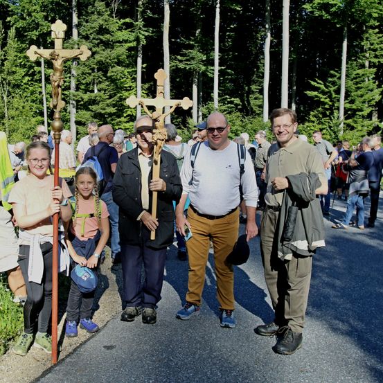 Vier Personen, zwei Frauen und zwei Männer, stehen mit Kreuzen und lächeln für ein Foto. Dahinter versammeln sich viele Menschen im Wald.