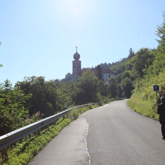 Eine Straße mit einem Leitplanken auf der linken Seite, einer Person, die geht, und einer Kirche mit einer Kuppel und einem Kreuz auf einem Hügel.