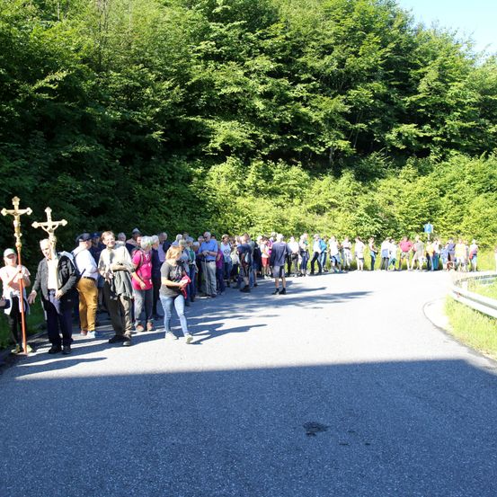Eine Gruppe von Menschen steht am Straßenrand, einige halten religiöse Kreuze. Sie sind von Grün und einem Waldhintergrund umgeben.