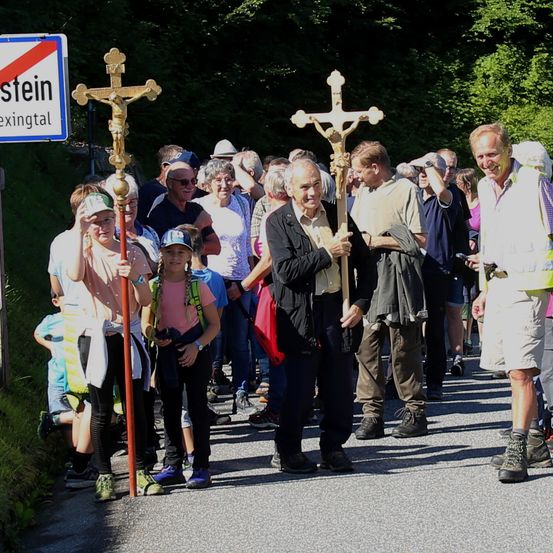 Eine Menschenmenge in einer Außenumgebung mit Kreuzen. Einige tragen Hüte, Brillen, Turnschuhe und Uhren. Ein Schild mit rotem Kreuz ist links angebracht.