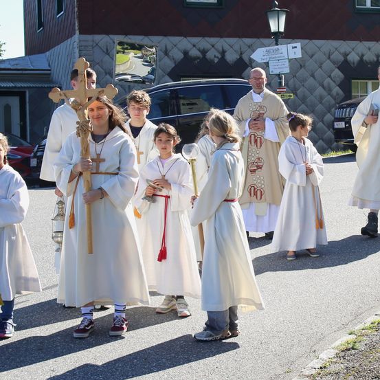 Eine Gruppe von Menschen in weißen Gewändern, wahrscheinlich während einer religiösen Prozession, geht auf der Straße. Eine Frau hält ein Holzkreuz und eine Laterne. Dahinter führt ein Priester die Prozession an.
