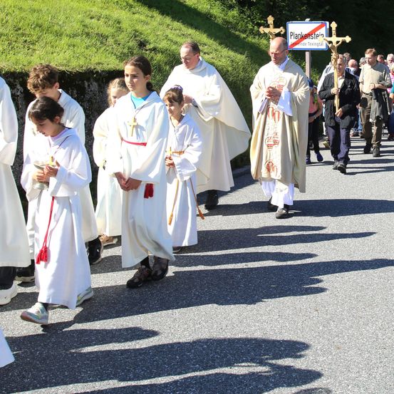 Eine Gruppe von Menschen in weißen Gewändern geht in einer Prozession, hält Kerzen und Kreuze. Ein Priester führt die Prozession an, mit einem Schild dahinter.