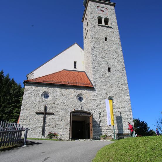 Eine steinerne Kirche mit einem hohen Turm und einem roten Dach, mit einem großen Kreuz und einer Fahne davor. Eine Person geht auf dem Rasen.