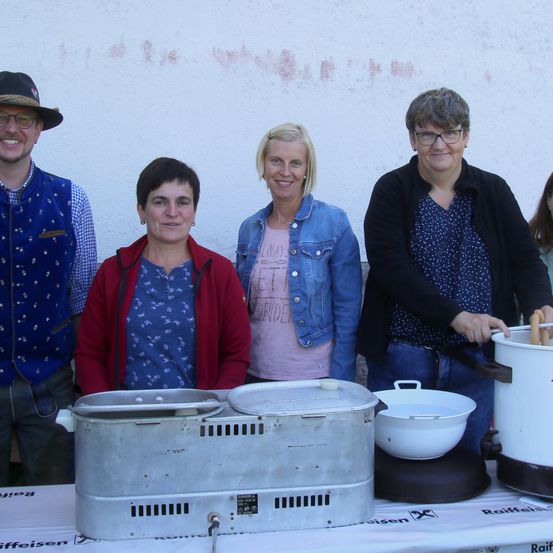 Fünf Personen stehen hinter einem Tisch mit Kochutensilien. Drei Frauen und ein Mann lächeln in die Kamera. Der Mann trägt einen Hut und eine blaue Weste. Der Tisch hat einen Hot-Dog-Dampfgarer und einen Wasserkocher.