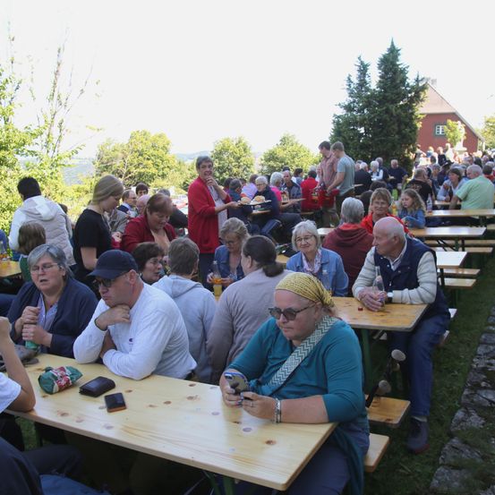 Eine große Gruppe von Menschen ist draußen versammelt und sitzt um Holztische herum. Einige unterhalten sich, während andere essen. Einige Leute tragen Brillen und Hüte. Bäume und ein Gebäude sind im Hintergrund.