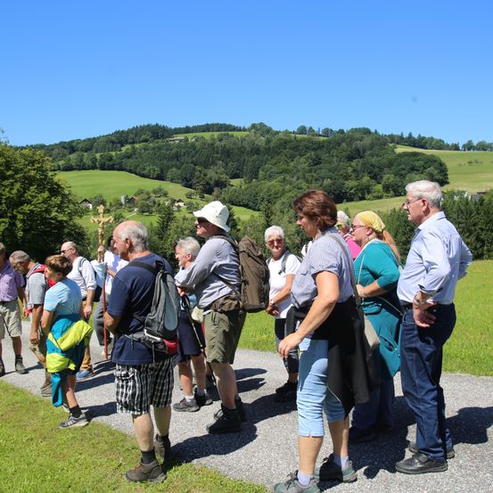 Eine Gruppe von Menschen, möglicherweise auf einer Wanderung, steht auf einem Weg in einem Grasfeld. Sie tragen Hüte, Brillen und Turnschuhe. Der Mann links hat einen Rucksack und einen Stock. Die Frau vorne hält eine Tasche. In der Ferne steht ein Kreuz und ein Berg.