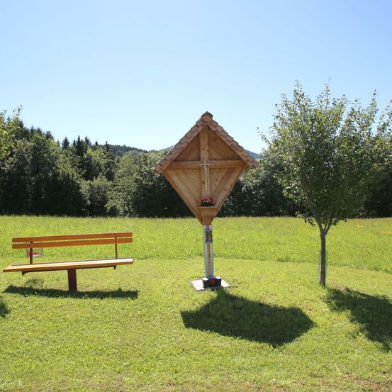 Eine Holzbank und eine kleine Holzkapelle stehen auf einer Rasenfläche mit einem Baum und Bergen im Hintergrund.