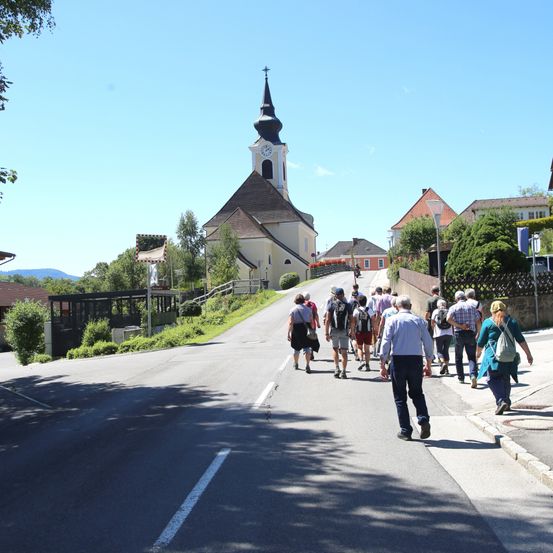 Eine Gruppe von Menschen geht vor einer Kirche mit einem Turm die Straße entlang. Die Kirche hat eine Uhr auf ihrem Turm. Sie tragen alle Rucksäcke und einige tragen Hüte. Auf der rechten Straßenseite befinden sich Häuser und Pflanzen. Auf der linken Seite gibt es Büsche und ein Gebäude.