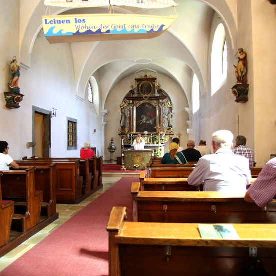 Das Innere einer Kirche mit hölzernen Kirchenbänken, einem Priester am Altar und einem Banner über dem Eingang. Die Leute sitzen in Reihen, und es gibt Statuen an den Wänden.