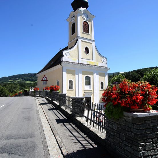 Eine kleine Kirche mit Glockenturm steht neben einer Straße, geschmückt mit roten Blumen und umgeben von Grün unter einem klaren Himmel.