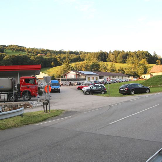 Ein Parkplatz mit einer Tankstelle, einem roten Lkw und Autos, die auf beiden Seiten der Straße geparkt sind. Die Landschaft zeichnet sich durch Hügel, Häuser und viel Grün aus.