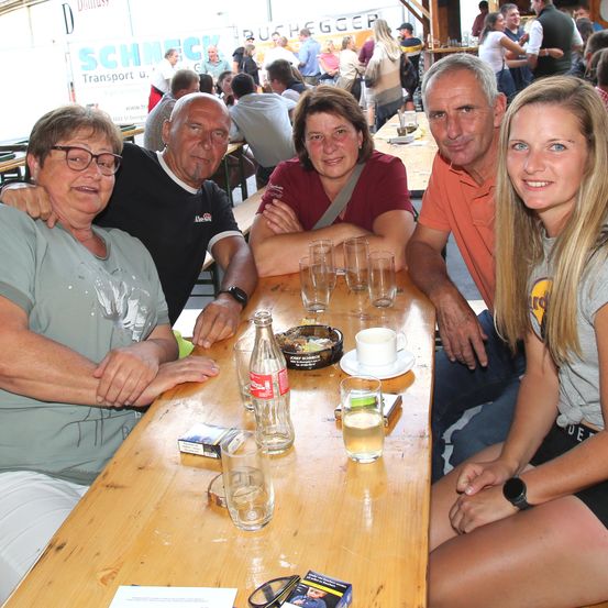 Fünf Menschen sitzen an einem langen Tisch, lächeln und haben eine Kulisse eines Outdoor-Events im Hintergrund. Verschiedene Gegenstände wie Gläser, eine Cola-Flasche und Zigaretten liegen auf dem Tisch.