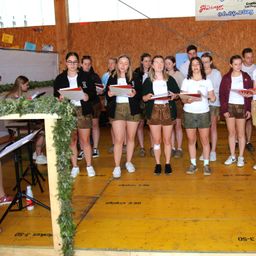 Eine Gruppe junger Menschen, sowohl Jungen als auch Mädchen, steht auf einer Bühne und singt mit Musikblättern in den Händen. Sie sind in traditioneller Kleidung gekleidet, und unter ihren Füßen befinden sich Holzplanken. Ein Banner und einige Texte befinden sich an der Wand hinter ihnen.