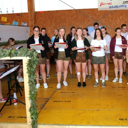 Eine Gruppe junger Menschen, sowohl Jungen als auch Mädchen, steht auf einer Bühne und singt mit Musikblättern in den Händen. Sie sind in traditioneller Kleidung gekleidet, und unter ihren Füßen befinden sich Holzplanken. Ein Banner und einige Texte befinden sich an der Wand hinter ihnen.