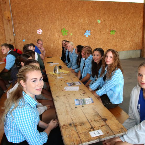 Eine Gruppe von Schülern sitzt an einem langen Holztisch und lächelt in die Kamera. Sie tragen passende blaue und weiße karierte Hemden. Die Wand hinter ihnen ist mit Holzpaneelen und bunten Blumen dekoriert.