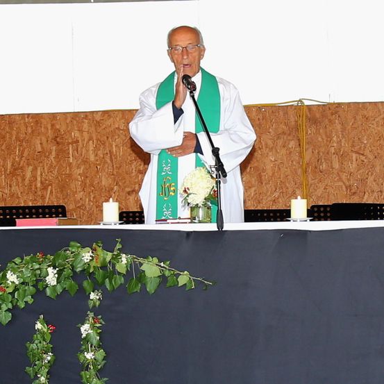 Ein älterer Priester steht an einem Podium und hält eine Rede. Er trägt eine weiße Robe mit einem grünen Schal und eine Brille. Zwei Kerzen flankieren eine weiße Blumenarrangement.