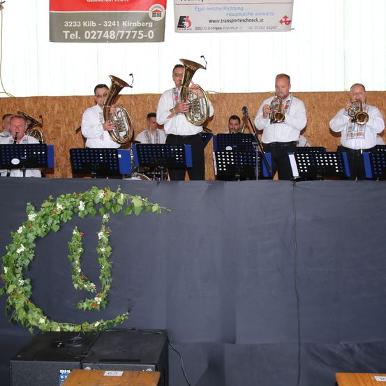 Eine Gruppe von Musikern spielt auf der Bühne mit verschiedenen Instrumenten. Die Bühne ist mit einem grünen Kranz geschmückt. Ein Banner mit der Adresse und Telefonnummer von Transportgeschneck hängt im Hintergrund.
