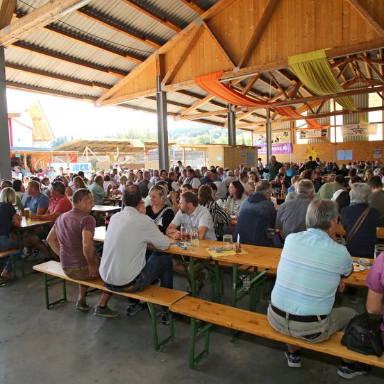 Eine große Menschenmenge versammelt sich in einem Pavillon mit Holztischen und -bänken, trinkt und unterhält sich. Der Pavillon hat Holzbalken und eine hohe Decke.